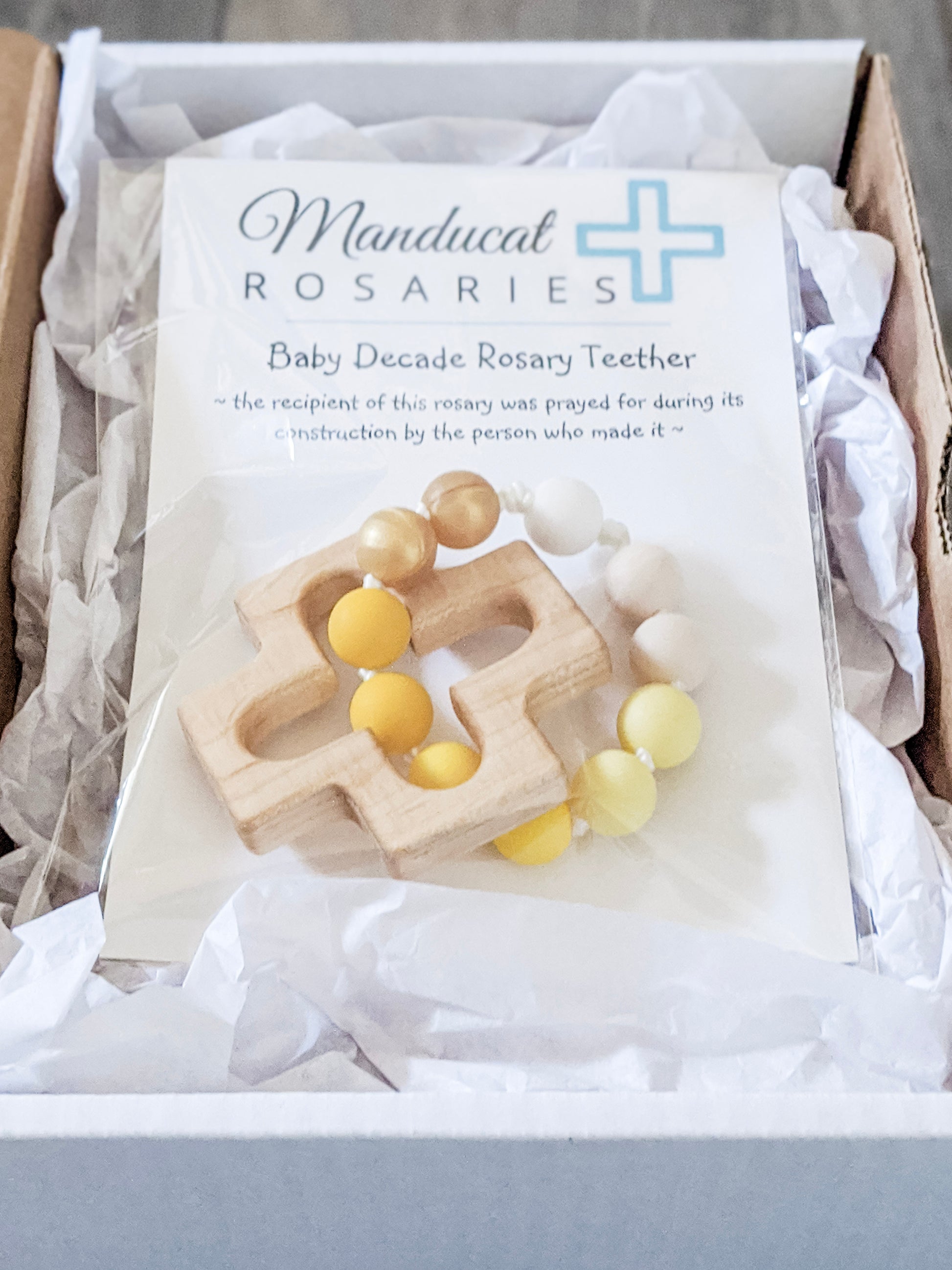 Yellow Catholic Baptism gift teething ring in packaging stating baby was prayed for during item's construction